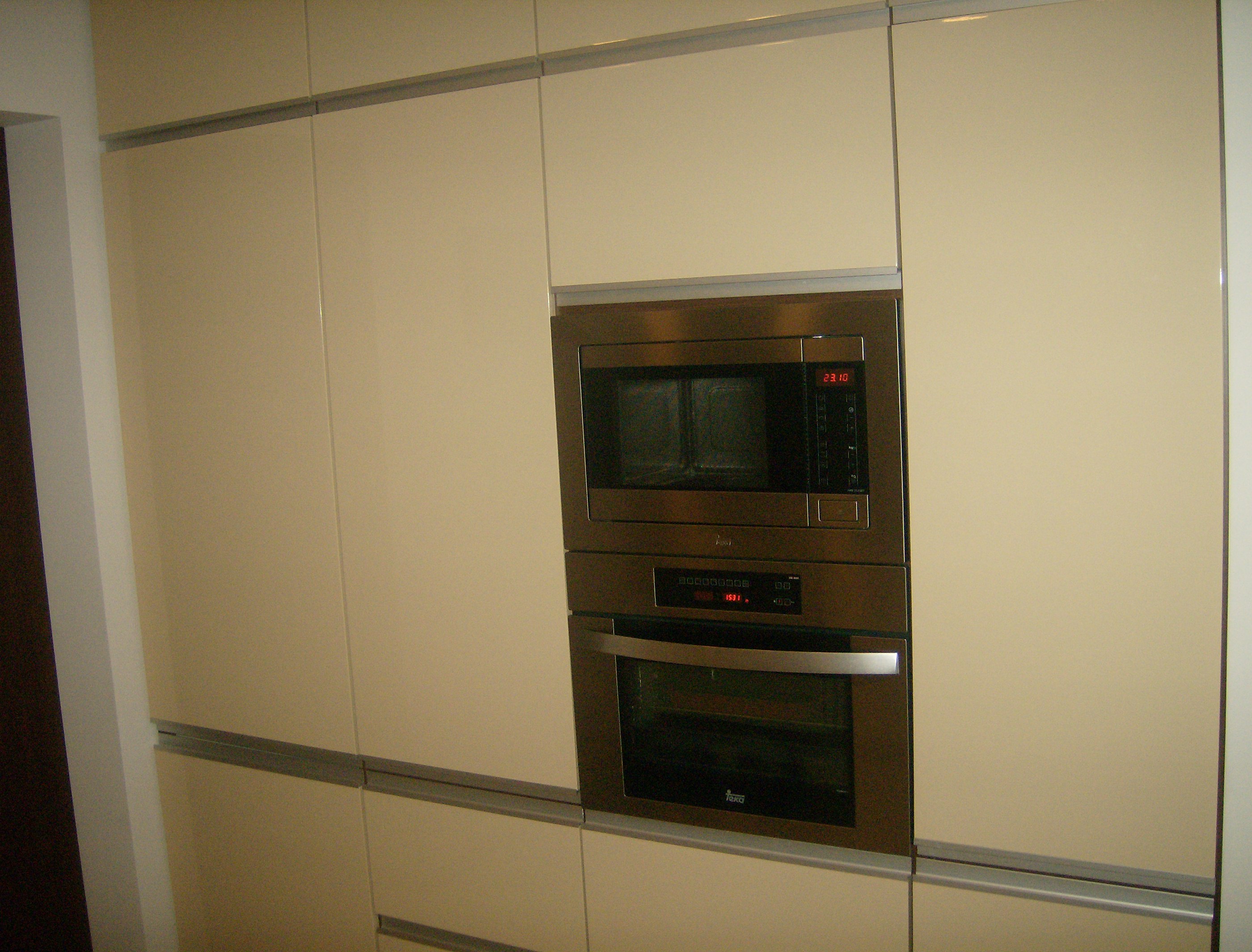 Beżowe, lakierowane fronty mebli kuchennych na wymiar z wbudowanym piekarnikiem i mikrofalówką w metalowej obudowie.